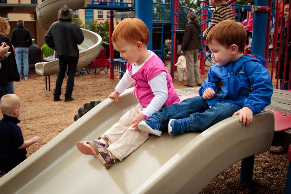 Two young children sitting at the top of a playground slide, ready to go down. Colorful playground equipment in the background.