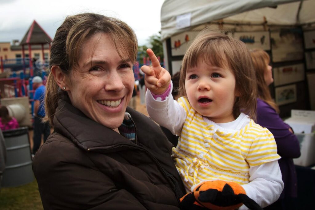 A woman smiles while holding a young child who points excitedly during a festival outing.