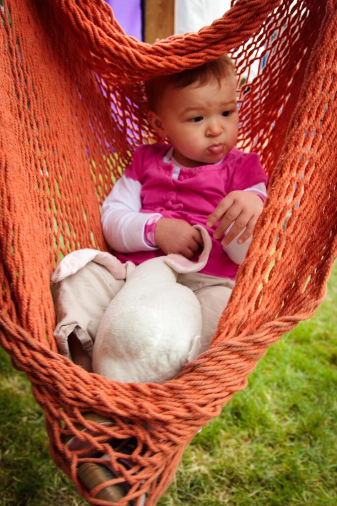 A toddler sits in an orange mesh hammock outdoors. The image includes a close-up of the hammock's woven texture.