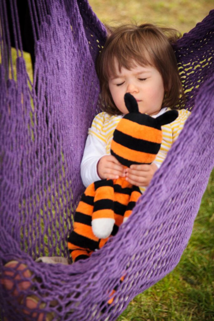 A child naps in a purple crocheted hammock while holding an orange and black striped bee stuffed animal.