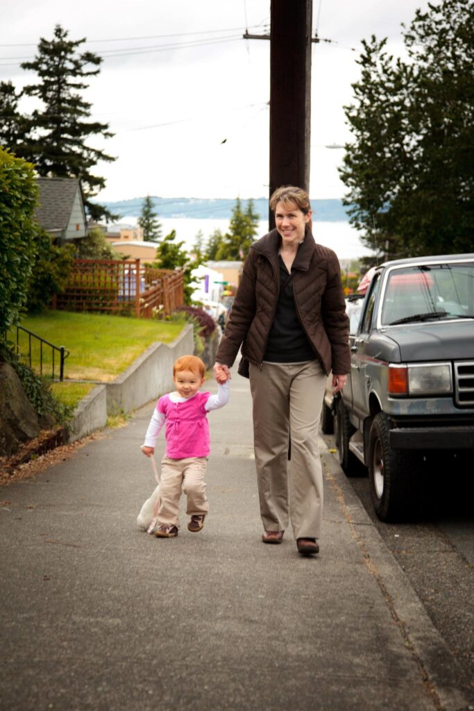 A family walks hand in hand on a residential street with a view of the ocean. They pass a parked truck and lush greenery.