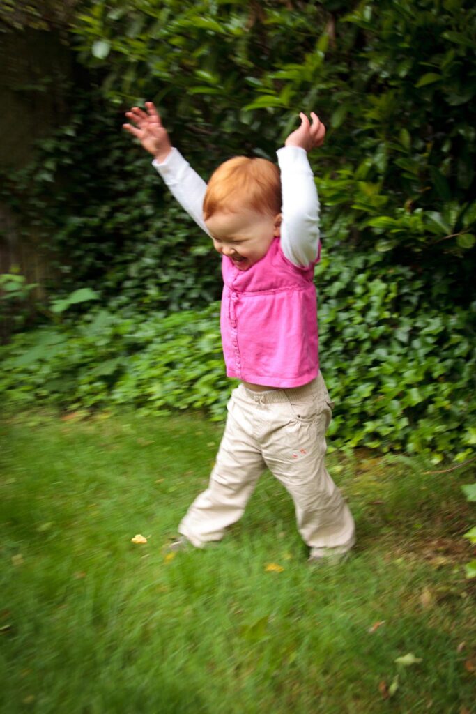 A toddler in a pink vest and beige pants dances on a grassy lawn. The background features lush green foliage.
