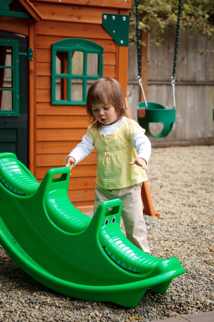 A child in a yellow vest plays on a green wooden rocker in the backyard.