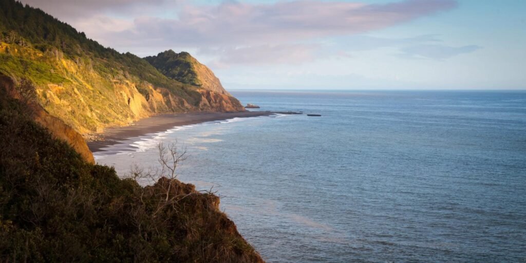 Dramatic view of a rugged Oregon coastline at sunset. Gentle waves roll onto a sandy beach bordered by lush, sunlit green hills.