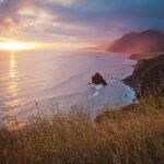 A serene sunset paints the Pacific Ocean in warm hues, viewed from a coastal hillside with rolling waves and rugged cliffs.