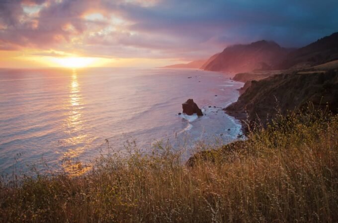 A serene sunset paints the Pacific Ocean in warm hues, viewed from a coastal hillside with rolling waves and rugged cliffs.