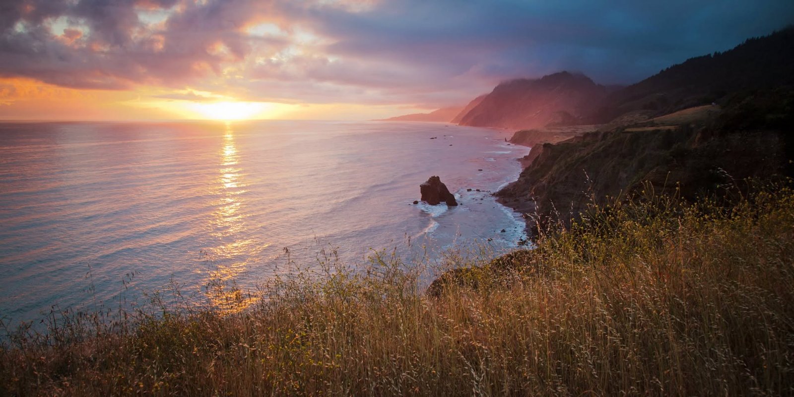 A serene sunset paints the Pacific Ocean in warm hues, viewed from a coastal hillside with rolling waves and rugged cliffs.