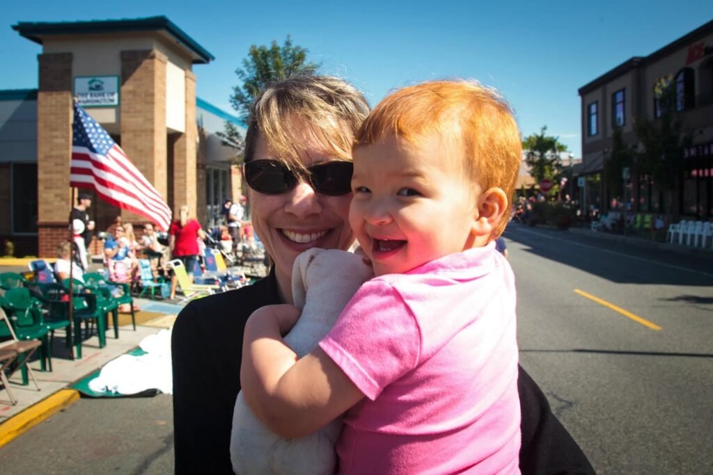 A woman and a red-haired toddler smiling together in an outdoor plaza. An American flag hangs from a building behind them on a sunny day.