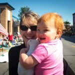 A woman and a red-haired toddler smiling together in an outdoor plaza. An American flag hangs from a building behind them on a sunny day.
