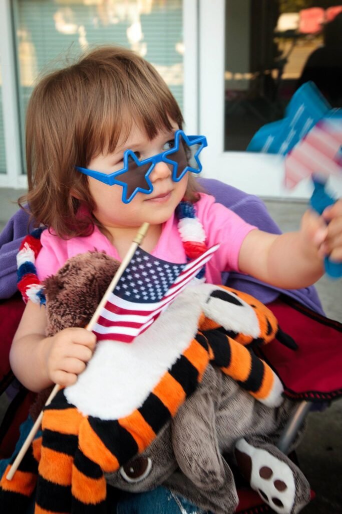 A child wearing blue star-shaped glasses and holding a small American flag at a parade.