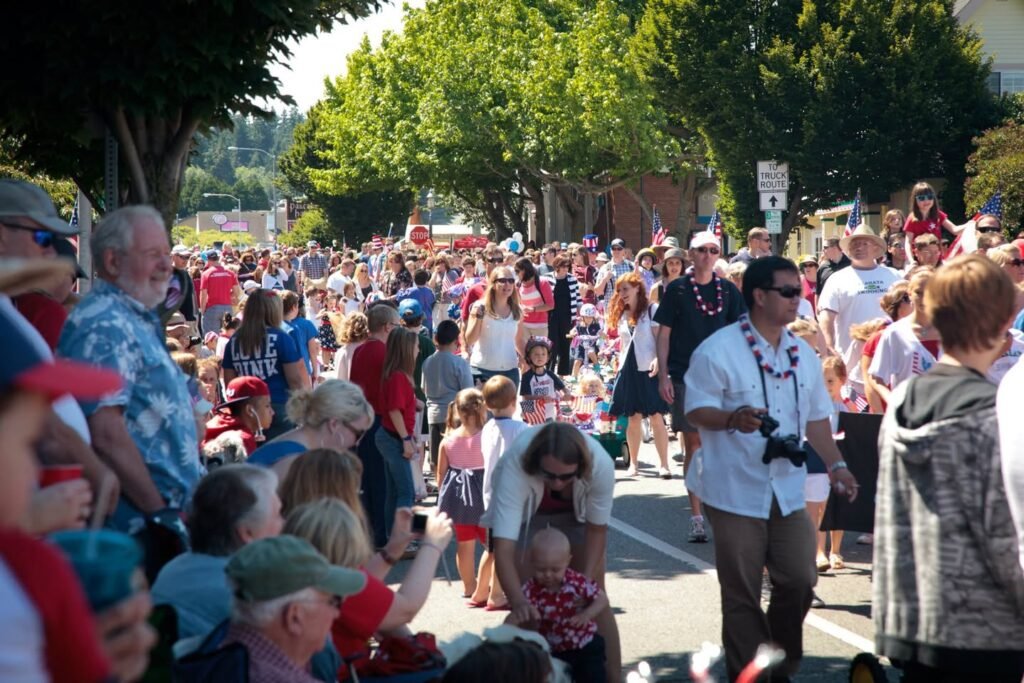 A vibrant community parade in the Pacific Northwest with people gathering on a sunny day. Crowds of diverse individuals watch from the sidelines surrounded by lush green trees.