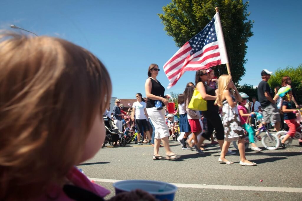 Candid view of a local parade with an American flag under a clear blue sky. People walk along a street lined with green trees.