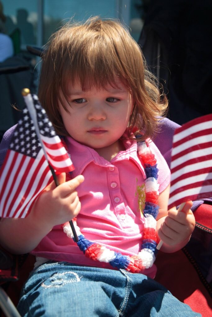 This image features a young child wearing a pink shirt and holding a small American flag. The child also has a red, white, and blue beaded lei around their neck, creating a festive patriotic atmosphere.