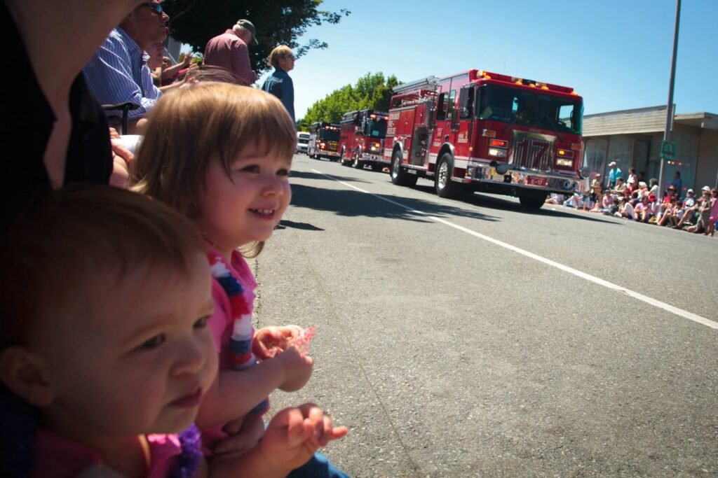 Children wave excitedly as a line of red fire trucks passes by during a sunny outdoor parade in the Pacific Northwest.