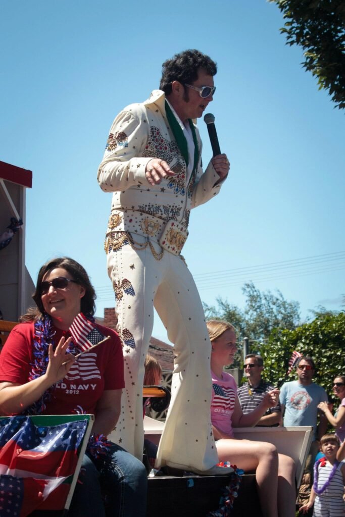 A performer dressed in a white sequined jumpsuit holds a microphone at a sunny outdoor festival.