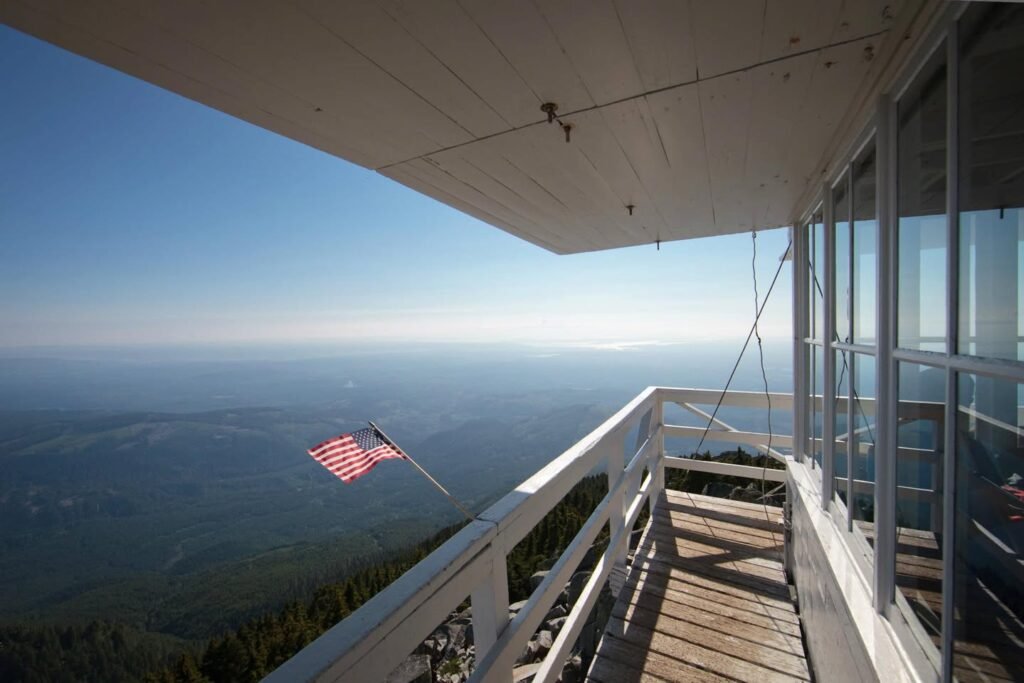 A wooden observation deck with white railings offers a panoramic view of a vast landscape below and an American flag flying in the wind under a clear blue sky.