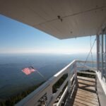 A wooden observation deck with white railings offers a panoramic view of a vast landscape below and an American flag flying in the wind under a clear blue sky.