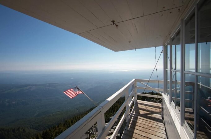 A wooden observation deck with white railings offers a panoramic view of a vast landscape below and an American flag flying in the wind under a clear blue sky.