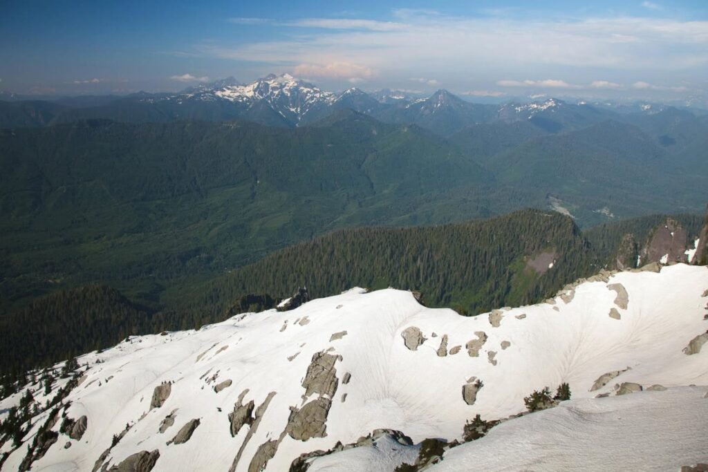 A high-angle view of a paved summit area overlooking deep green forested valleys. A vast mountain range stretches into the hazy distance under a clear sky.