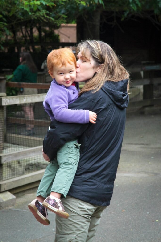 An adult kisses a red-haired child holding them up near a wildlife enclosure with trees in the background.