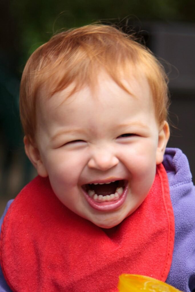 A joyful toddler laughing with a red scarf and purple coat outdoors.