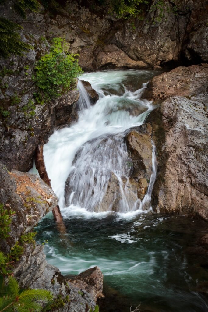 A powerful waterfall cascades over rugged rocks into a deep pool. Lush green vegetation surrounds the rocky cliffs.