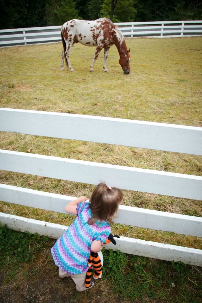 A child in a colorful striped dress stands by a white fence, observing a spotted horse grazing in a pasture.