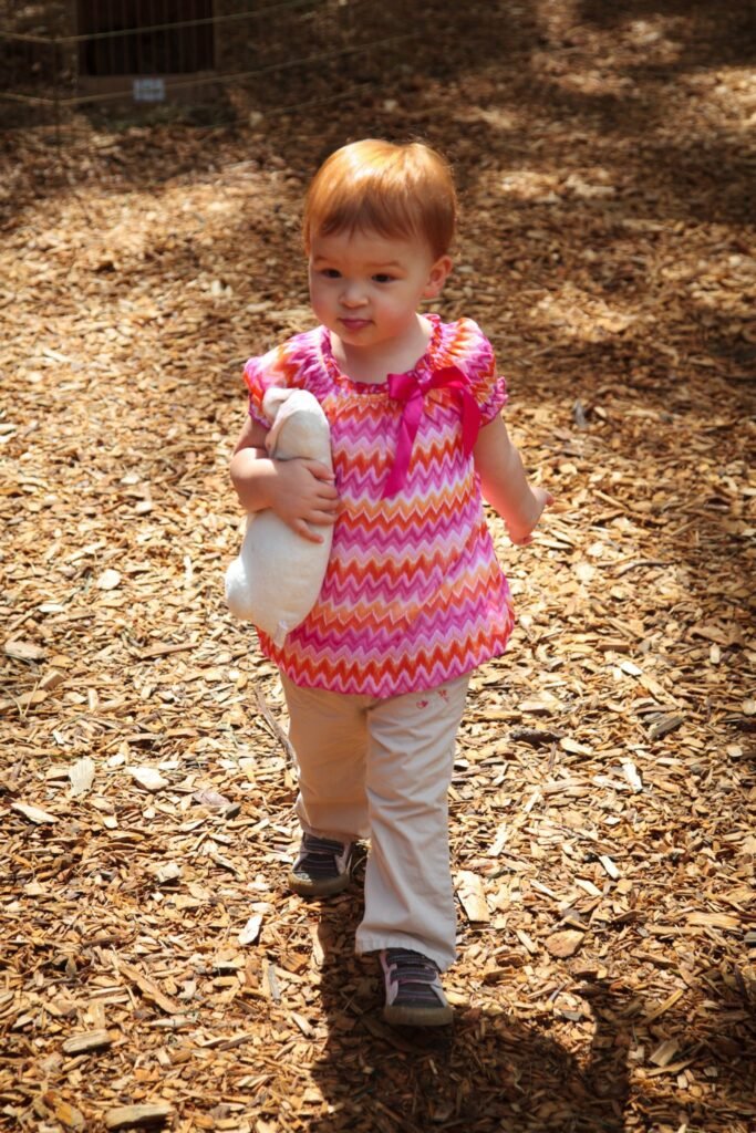 A young child holding a plush toy and walking over a layer of wood chips outdoors.