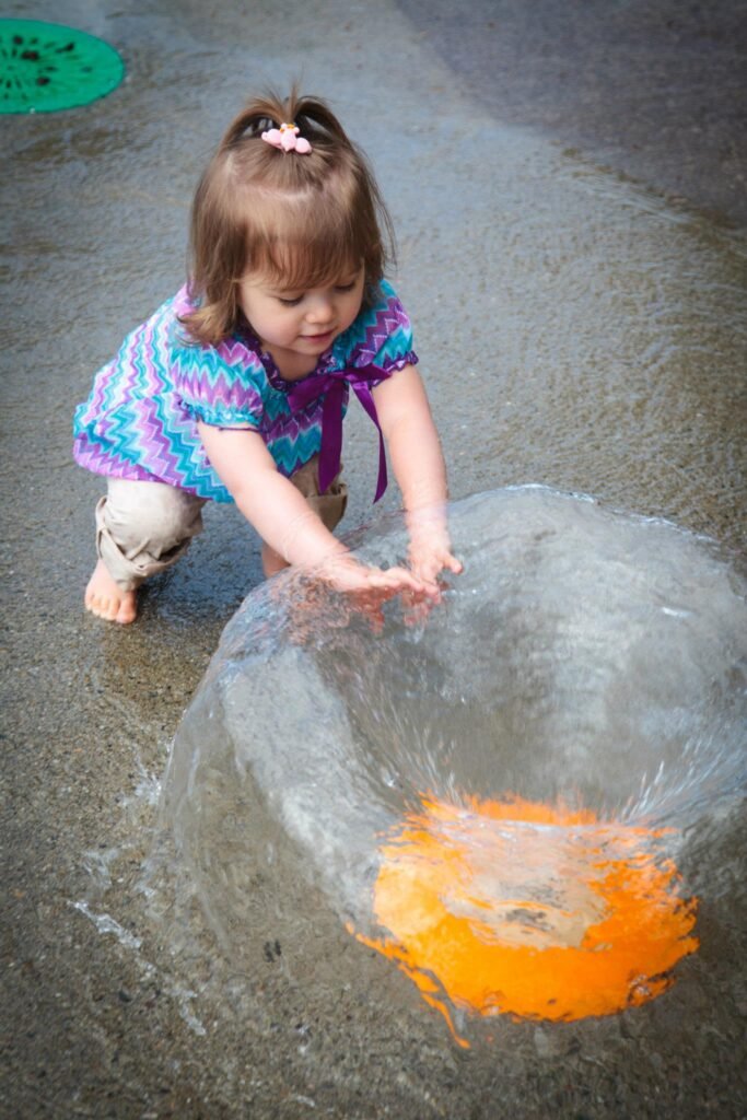 A young child plays with water at a fountain with bright orange accents on a paved surface.