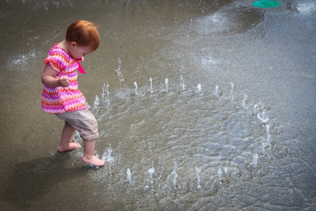 A child in a pink and white striped outfit walks through small water jets at a public fountain.