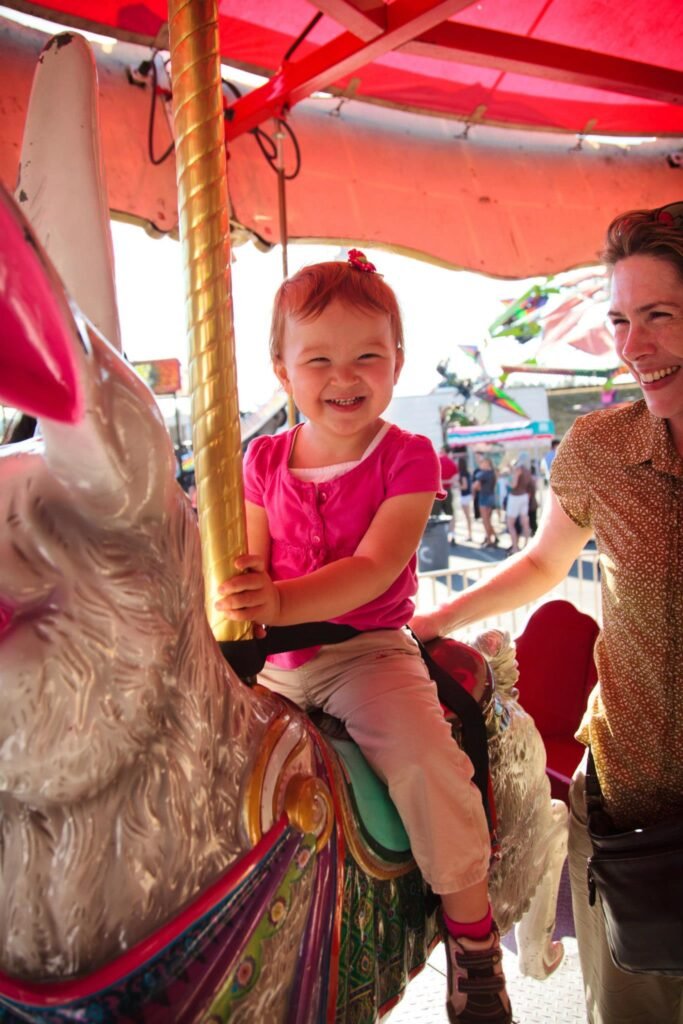 A child in a pink shirt rides a carousel horse with an adult beside them in a carnival setting.