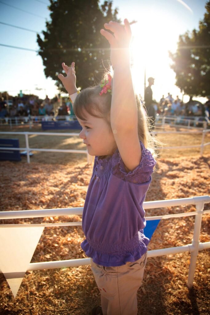 A young child in a purple shirt jumps with arms raised in a fenced-area covered in wood chips under bright sunlight.