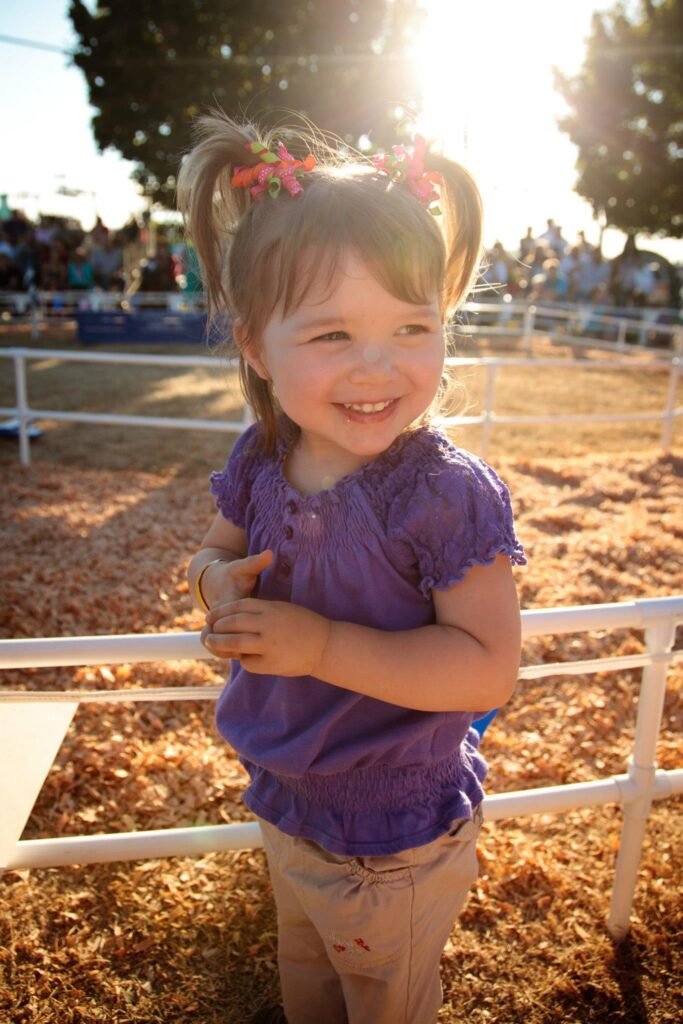 A young girl with pigtails smiles while holding a white fence at an outdoor event. The background features wood chips and other visitors.