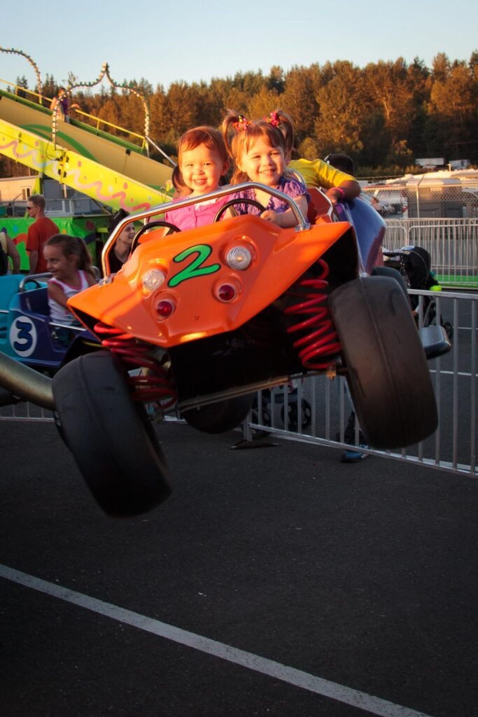 A small child ride-on vehicle labeled number 2, orange with big tires, is airborne on a track at a fairground on a clear evening with trees in the background.