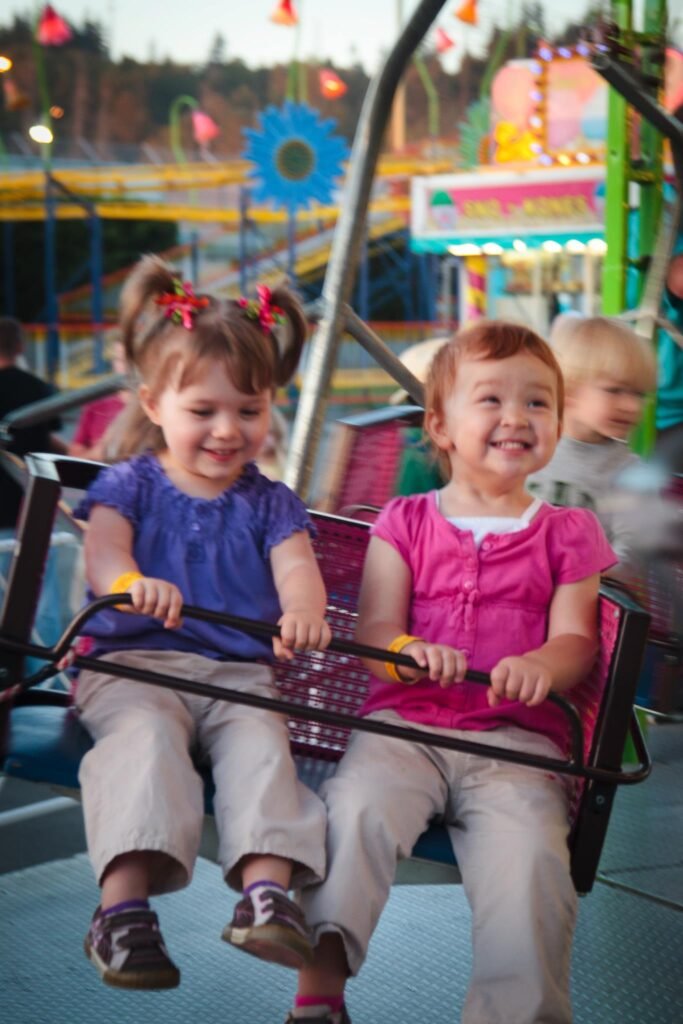 A young girl wearing a solid-pink short-sleeve shirt and beige trousers smiles from a carnival roller coaster. The colorful fairground background includes a sunflower decoration and stacked photo panels showing other kids in the same seat.
