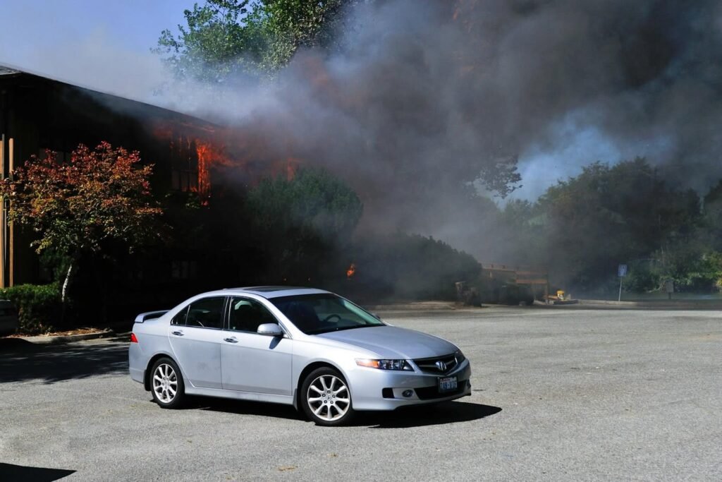 A silver sedan parked in an open lot with thick smoke and visible flames engulfing a building in the background.