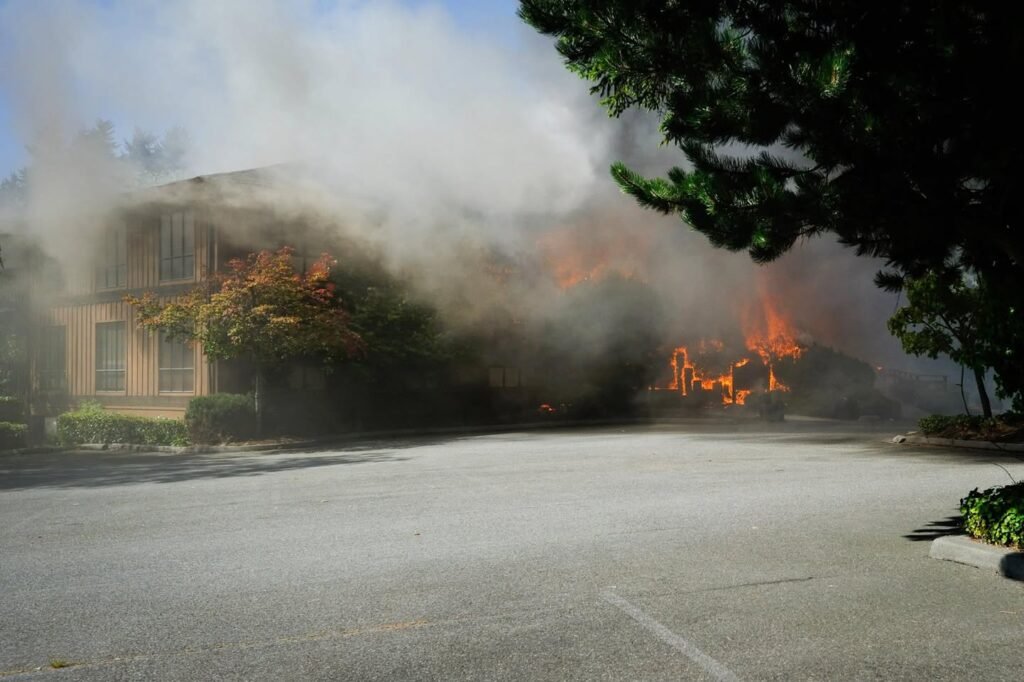 Flames and thick smoke rise from a fire near a house and trees in a paved area. This scene captures the intensity of recent wildfire activity in the region.