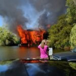 A striking scene of a burning car with intense flames and dark smoke rising from a green shoreline.