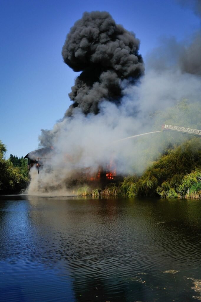 A large plume of black smoke rises from the shoreline of a lake, with firefighting water streams visible against a clear blue sky.