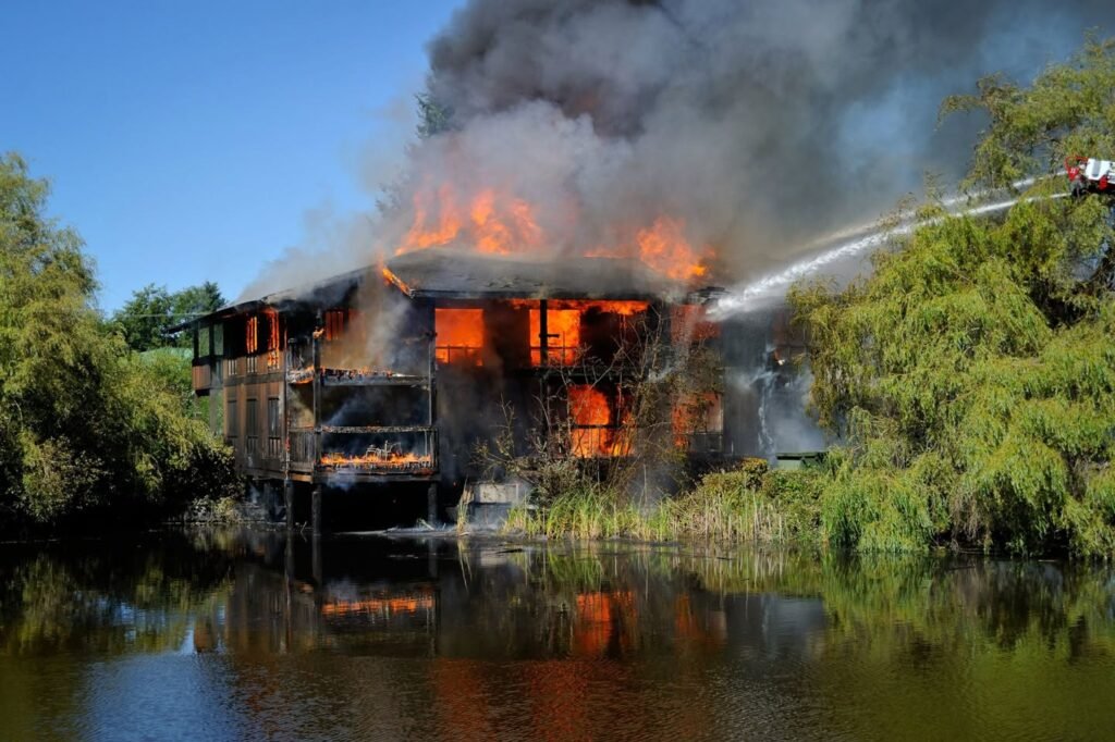 A house located on the water's edge is engulfed in flames with smoke rising while a high-pressure water jet is aimed at the burning building.