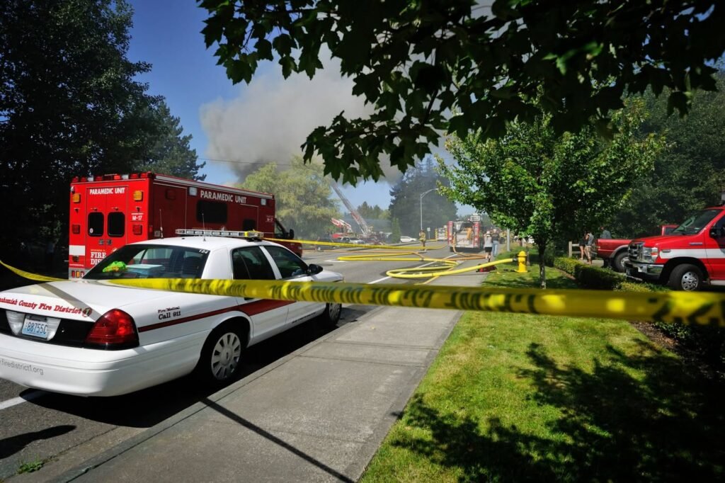 A police car and fire truck are on scene with yellow caution tape and smoke rising from the background.