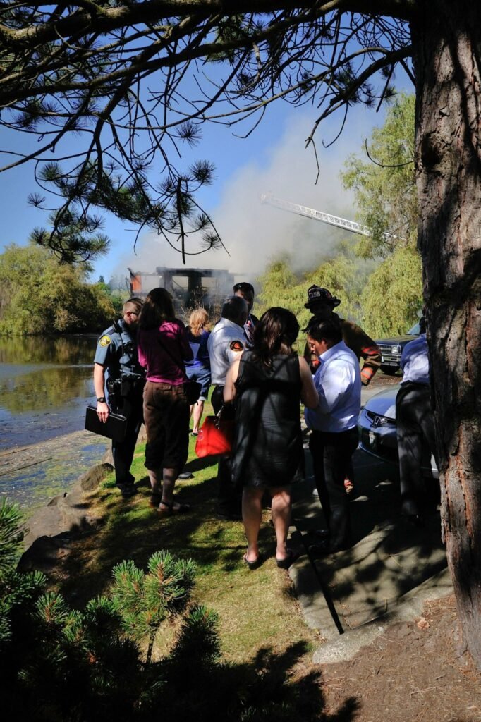 Visitors stand near a steaming structure at a lakeside in the Pacific Northwest under a clear sky.