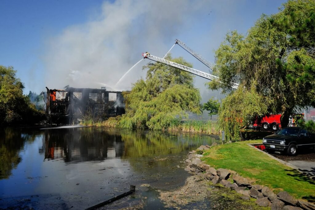 A lagoon-side structure is engulfed in smoke as a fire truck's aerial ladder sprays water to extinguish the blaze, with a red truck and black sedan nearby.