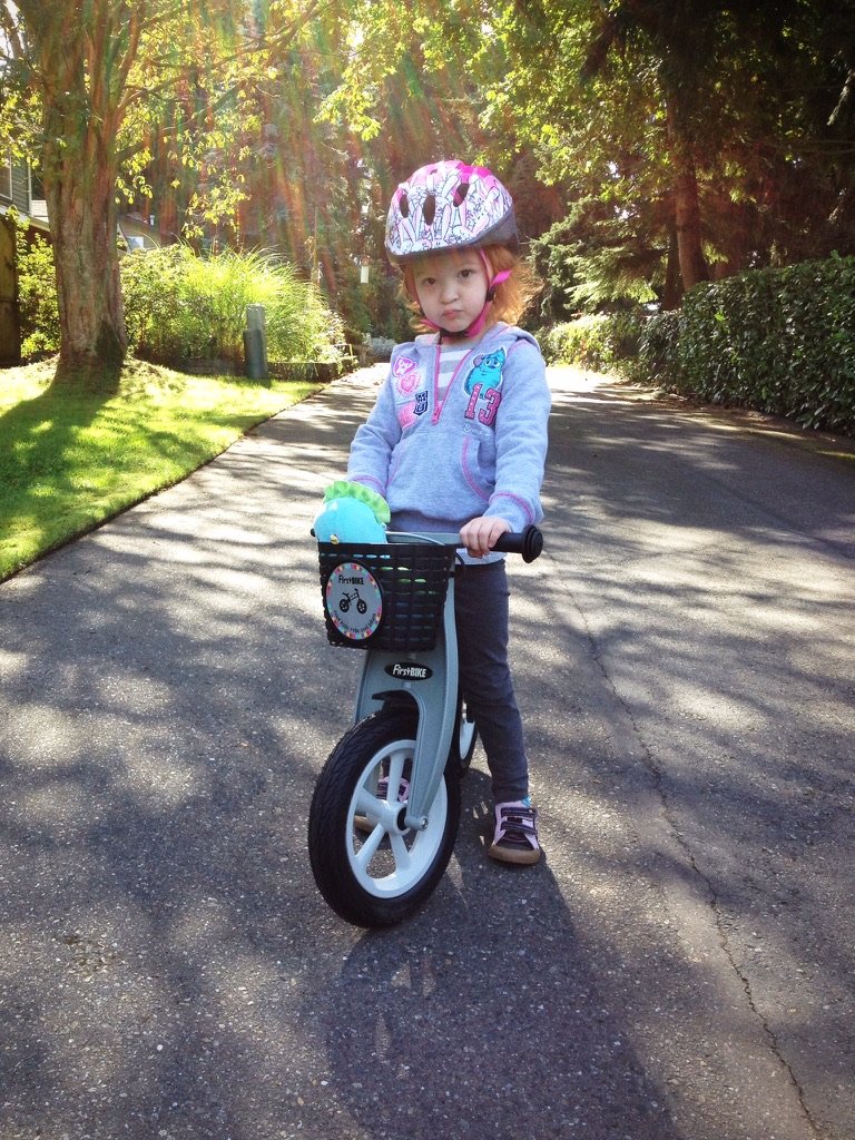 A child stands on a balance bike on a sunlit street. The background is a quiet residential road lined with trees.