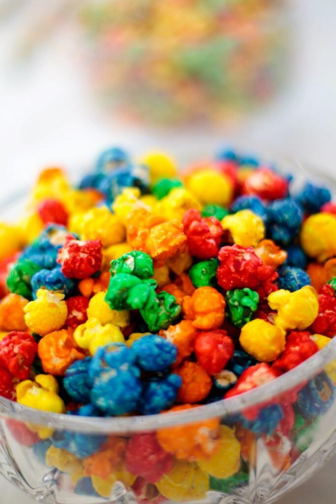 A vibrant glass bowl filled with rainbow-colored popcorn, surrounded by blurred background elements.