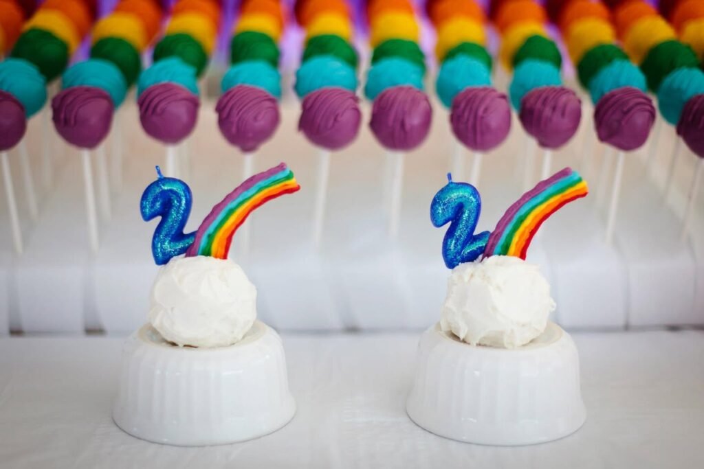 Colorful cakeshots decorated with white frosting, number two candles, and rainbow arcs on a white surface.