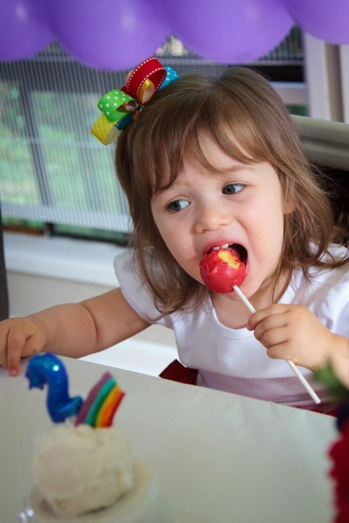 A child with a colorful hair accessory takes a big bite of a red lollipop at a birthday table.