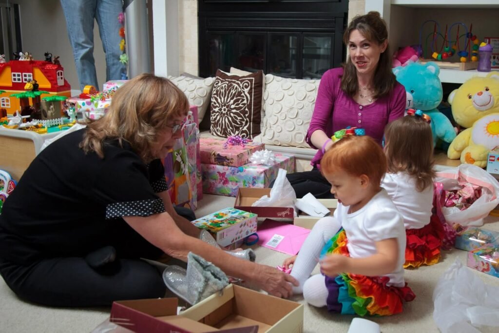 A family enjoys a cozy gathering on the living room floor, opening wraps together while seated near a sofa with colorful toys visible in the background.