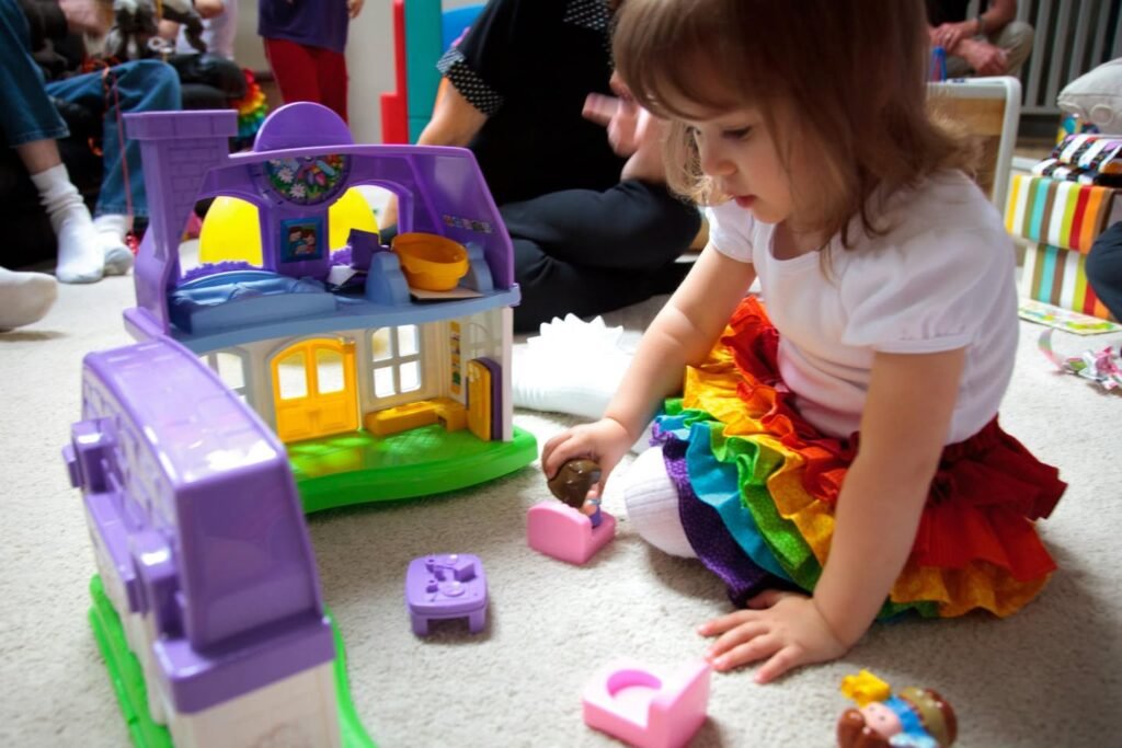 A child in a colorful rainbow skirt sits on the floor playing with a toy house and garden carts during a joyful family moment.