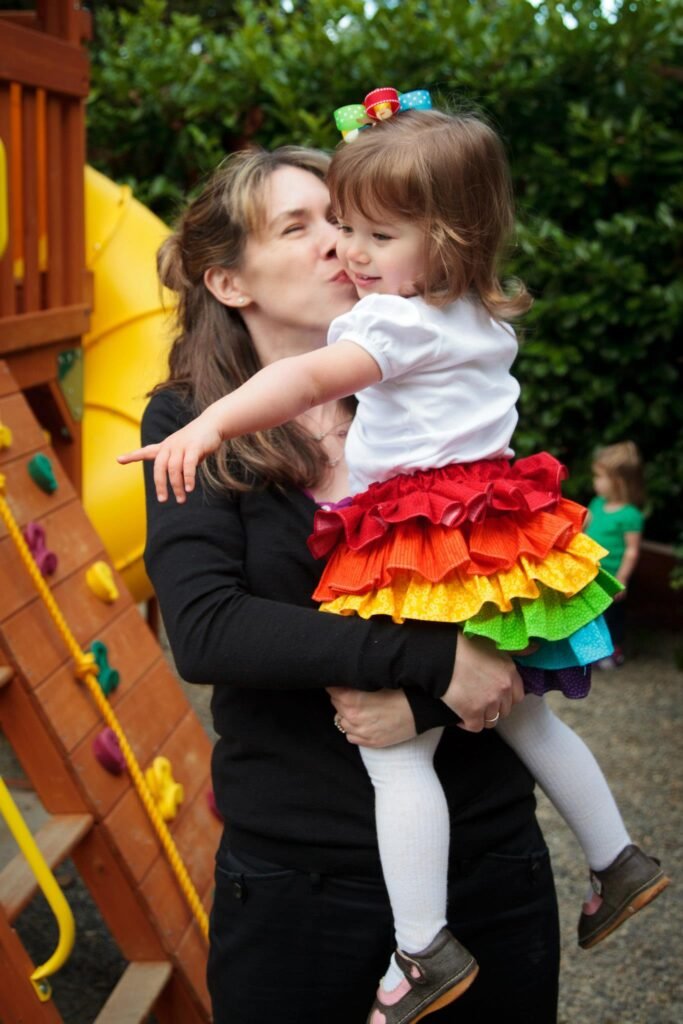 A person holding a child wearing a colorful rainbow skirt near a wooden playset.